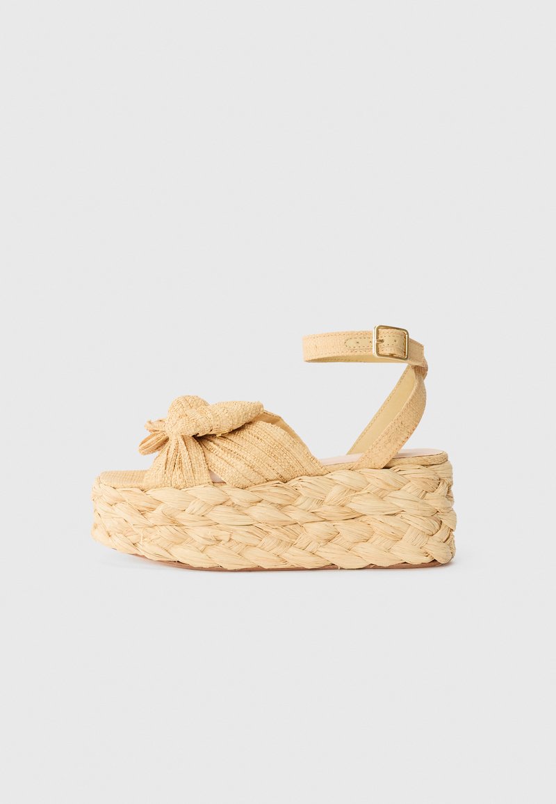 Platform sandals featuring a knotted design, woven straw base, and a light yellow fabric upper. Adjustable ankle strap with a gold buckle.