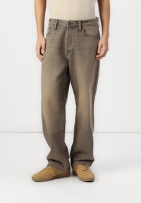 Jean en denim léger marron clair, ample et à la finition délavée, avec cinq poches et une fermeture à bouton classique, porté avec des baskets beiges.