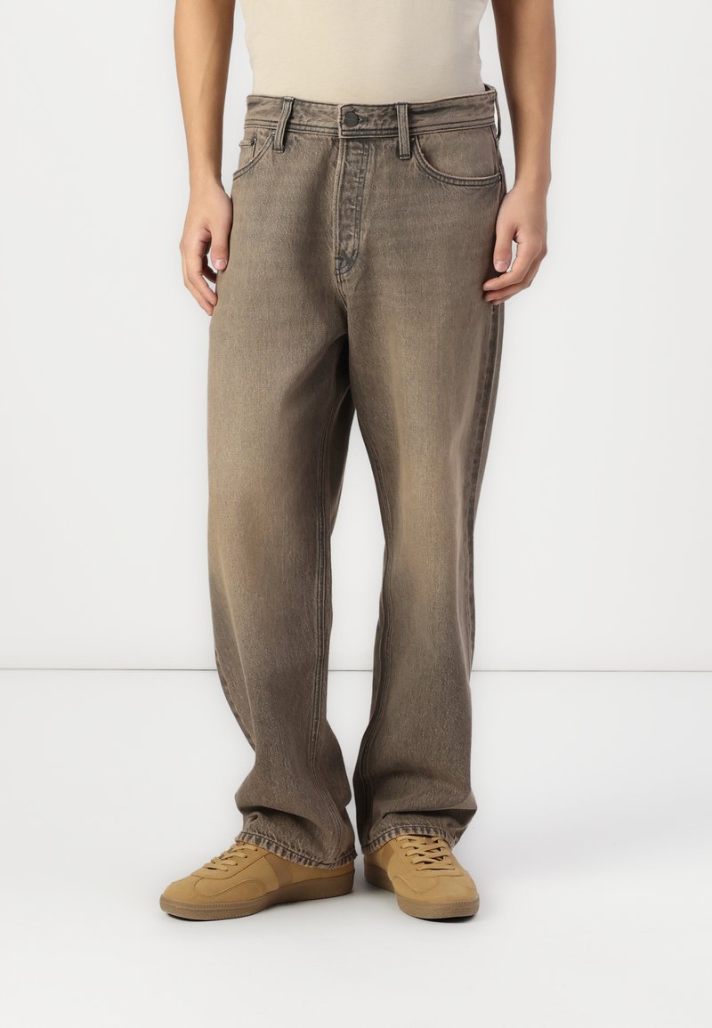 Jean en denim léger marron clair, ample et à la finition délavée, avec cinq poches et une fermeture à bouton classique, porté avec des baskets beiges.