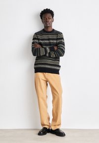 Schwarzer Strickpullover mit grünen und weißen geometrischen Mustern, kombiniert mit beigen Hosen und schwarzen Schuhen. Das Modell steht vor einem weißen Hintergrund.