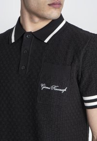 Černý texturovaný polo tričko s žebrovaným límcem s bílými pruhy, dvěma knoflíky a levým kapsičkou na hrudi s bílou výšivkou.