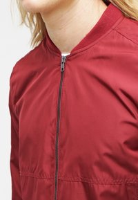 Giacca bomber rossa, materiale in nylon, chiusura con zip, colletto a coste, texture leggera, design minimalista, presenta tasche laterali.