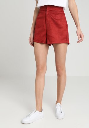 Shorts taille haute en daim rouge avec poches frontales, design ajusté et texture lisse, assortis à des baskets blanches.