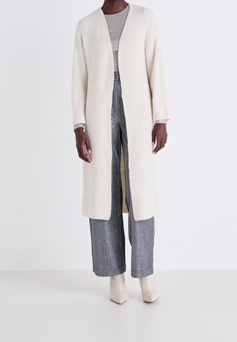 Personne portant un long cardigan crème ouvert sur le devant, un col roulé gris, un pantalon gris à jambes larges, et des bottines blanches cassées sur un fond uni.