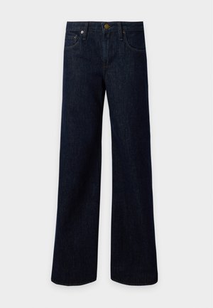 Kvinde stående med hænderne i lommerne iført sort militærinspireret jakke, sort top, mørke vidde jeans og sorte sko mod ensfarvet baggrund.