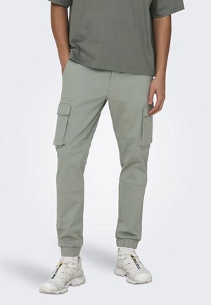 Homme portant un pantalon jogger cargo vert clair, un t-shirt vert olive et des baskets blanches à rayures multicolores, debout avec une main dans la poche.