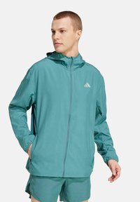Chaqueta deportiva en color teal con cremallera frontal, capucha y puños elásticos. Presenta un logo sutil en el pecho y bolsillos laterales. Tejido suave y ligero.