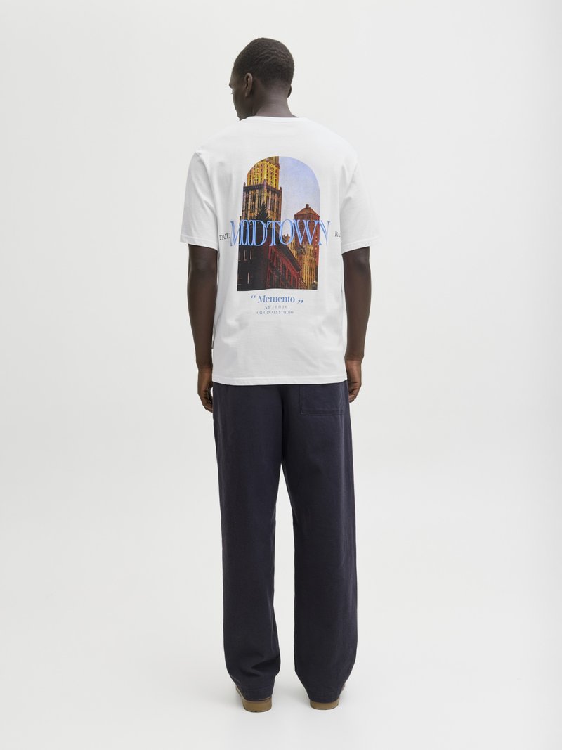 Muž v bielom tričku s mestskou krajinou a textom "MOODTOWN" na chrbte, v kombinácii s voľnými tmavými nohavicami, stojaci pred jednofarebným pozadím.