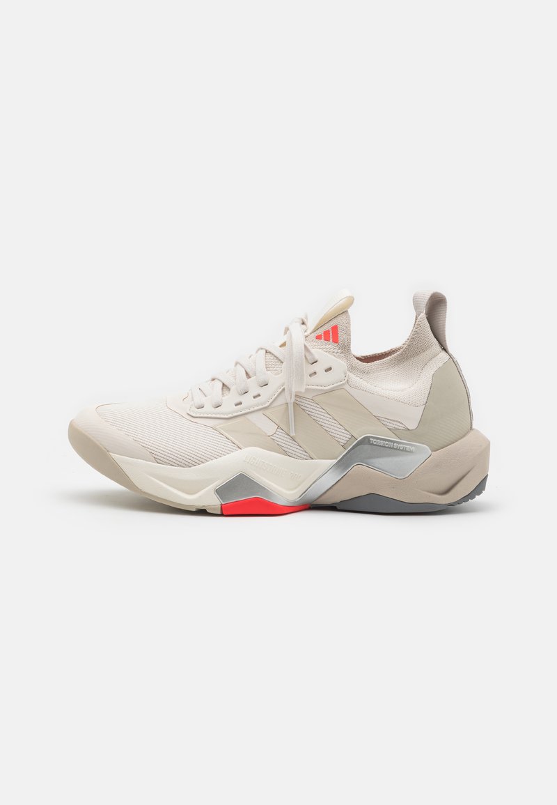 Light beige athletic shoe with a textured upper, three diagonal stripes, silver accents, and a red outsole detail. Features a chunky heel design.