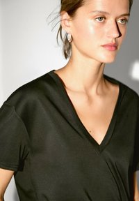 Czarna koszulka z dekoltem w serek, wykonana z miękkiego materiału, z krótkimi rękawami i luźnym krojem. Modelka nosi srebrne kolczyki, ma gładką skórę i delikatne oświetlenie.