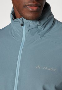 Veste zippée bleu clair en tissu lisse. Dotée d'un col haut, d'une fermeture éclair sur le devant, et d'un petit logo gris sur la poitrine.