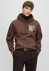 Hoodie marron avec une poche avant, orné d'un patch carré de fleurs jaunes sur un fond gris, capuche avec cordon de serrage et poignets côtelés.