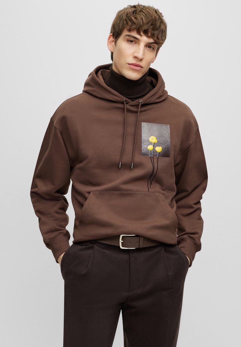 Hoodie marron avec une poche avant, orné d'un patch carré de fleurs jaunes sur un fond gris, capuche avec cordon de serrage et poignets côtelés.