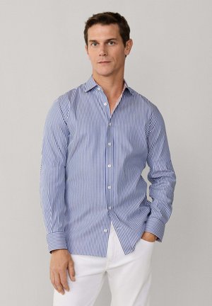 Homme portant une chemise à rayures verticales bleues et blanches et un pantalon blanc, debout devant un fond gris clair uni.