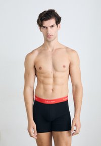 Boxer in cotone nero con vita elastica rossa, con scritta "Calvin Klein" stampata in bianco, modello con un fisico tonico, posato frontalmente.