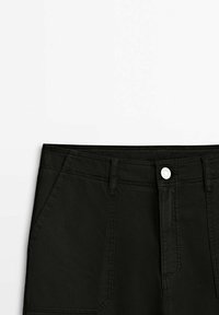 Mustapukuiset farkut, joissa on tasainen vyötärö, etupocketit ja metallinen napitus. Kangas on sileäpintainen, ja siinä on hienovaraisia ompeluyksityiskohtia.