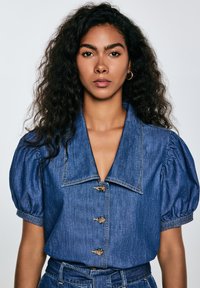 Denimshirt med stor krage, volangermer och kantknäppning. Har en strukturerad blått tyg och guldtonade knappar.