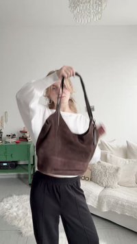 Person i vit topp och svarta byxor som håller upp en mörkbrun handväska i ett ljust vardagsrum med ett grönt skåp och en vit soffa.