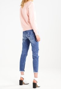 Sudadera de algodón rosa, jeans de mezclilla azul desgastados y sandalias de tacón bloque blanco. Los jeans tienen dobladillos deshilachados y un corte relajado.