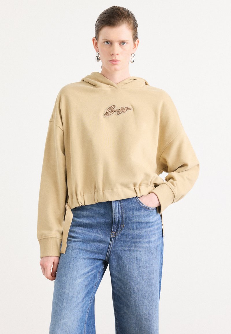 Beige cropped hettegenser med snor og brodert tekst. Stoffet har en myk tekstur, sammen med lyseblå høytlivsjeans.