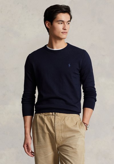 Jonge man die een donkerblauwe trui en beige broek draagt, kijkt zijwaarts met een hand in zijn zak tegen een effen achtergrond.
