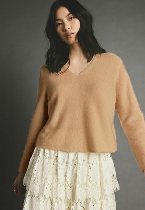 V-Ausschnitt, langärmeliger Pullover aus weichem beigem Stoff, kombiniert mit einem mehrlagigen weißen Spitzenrock mit floralen Mustern. Texturierter Kontrast in den Stoffen.