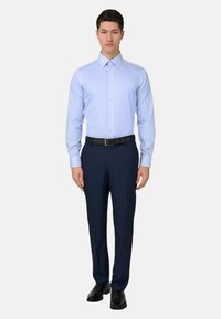 Mann steht, trägt hellblaues Hemd, dunkelblaue Anzughose, schwarzen Gürtel und schwarze Lederschuhe vor einfachem Hintergrund.