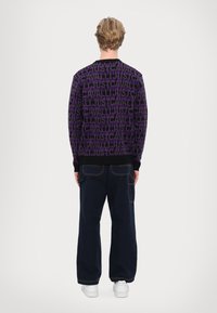 Purple sweater with repeating text pattern, long sleeves, and black ribbed collar. Paired with navy wide-leg pants and white sneakers.