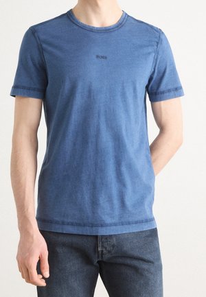 Mand iført en tætsiddende blå T-shirt med korte ærmer med "BOSS" logo på brystet og mørkeblå jeans foran en ensfarvet baggrund.