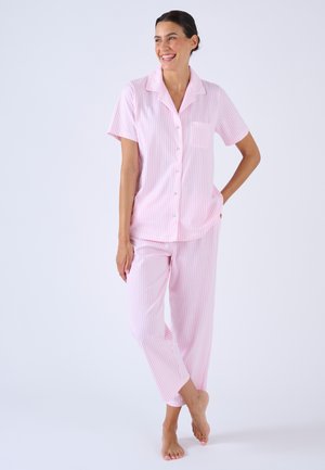 Vrouw staat blootsvoets en draagt een roze-wit gestreept pyjamapak met korte mouwen en bijpassende lange broek, ze lacht en kijkt naar rechts.