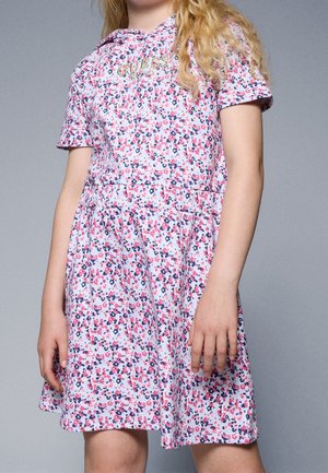 Mädchen trägt ein kurzärmeliges Kleid mit pinken und marineblauen Blumenmuster und dem goldfarbenen "GUESS"-Logo auf der Brust, steht vor einem grauen Hintergrund.