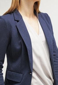 Veste bleu marine à la texture lisse, fermeture à un bouton et coupe structurée, associée à un chemisier blanc drapé en dessous.