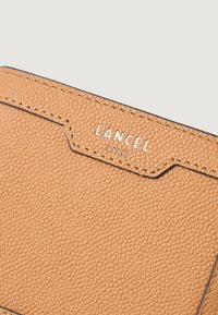Portfel z teksturowanej skóry w kolorze brązowym z wytłoczonym logo "Lancel Paris", charakteryzujący się kontrastowym ciemnym przeszyciem i eleganckim, kanciastym kształtem.