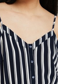 Top a righe blu navy e bianche con spalline sottili, scollatura a V e dettagli con bottoni sul davanti. Tessuto liscio con linee verticali.