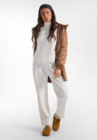 Bruine faux suede jas met capuchon, witte geribde trui, witte corduroy broek en tan slippers. Warme texturen en neutrale kleuren worden getoond.