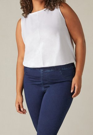 Vrouw met krullend haar die een mouwloos wit shirt draagt, ingestopt in een high-waisted donkerblauwe spijkerbroek, tegen een effen achtergrond.