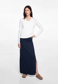 Bijela bluza s dugim rukavima u kombinaciji s tamnoplavom maksi suknjom s bočnim prorezom, izrađena od mekanog materijala, upotpunjena smeškim slip-on cipelama.
