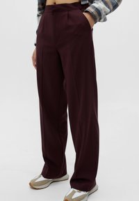 Personne portant un pantalon large taille haute de couleur bordeaux foncé avec poches latérales, assorti à des baskets beige et blanches sur un fond uni.