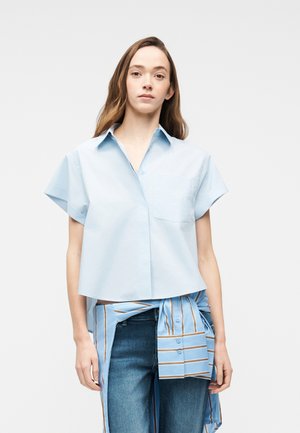 Junge Frau mit langen Haaren trägt ein lockeres, hellblaues Kurzarmhemd und blaue Jeans, mit einem gestreiften Hemd, das um die Taille gebunden ist.