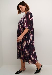 Robe fluide, mi-longue, avec un motif abstrait violet et noir, dotée de manches courtes et d'un tissu léger. Associée à des chaussures noires.