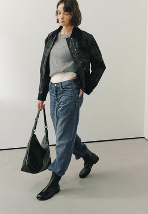 Kvinde går indenfor iført en sort ternet jakke, grå sweater, blå jeans, sorte støvler og bærer en sort håndtaske.