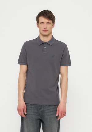 Jauns vīrietis ar īsiem brūniem matiem, valkājot tumši pelēku īsroku polo kreklu un izbalējušas pelēkas džinsas, stāvot pret baltu fonu.