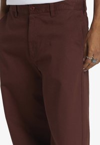 Burgunderfarbene Baumwollhosen verfügen über einen Bund mit Knopfverschluss, seitliche Taschen und ein gerades Bein-Design, das eine glatte Textur zur Schau stellt.