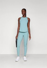 Débardeur de sport bleu clair et leggings assortis, en tissu stretch. Le modèle tient une bouteille d'eau noire et porte des chaussures de sport blanches.
