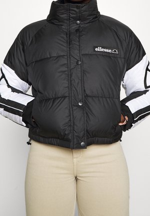 Schwarze Steppjacke mit weißen und schwarzen Ärmeln, Druckknöpfen und ellesse-Logo auf der Brust, getragen mit beigen Hosen und Händen in den Taschen.