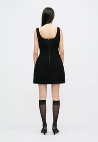 Μαύρο αμάνικο φόρεμα με εφαρμοστό κορσάζ, φουσκωτή φούστα και φερμουάρ στην πλάτη. Το μοντέλο φοράει διάφανα μαύρα καλσόν μέχρι το γόνατο και ψηλοτάκουνα παπούτσια.