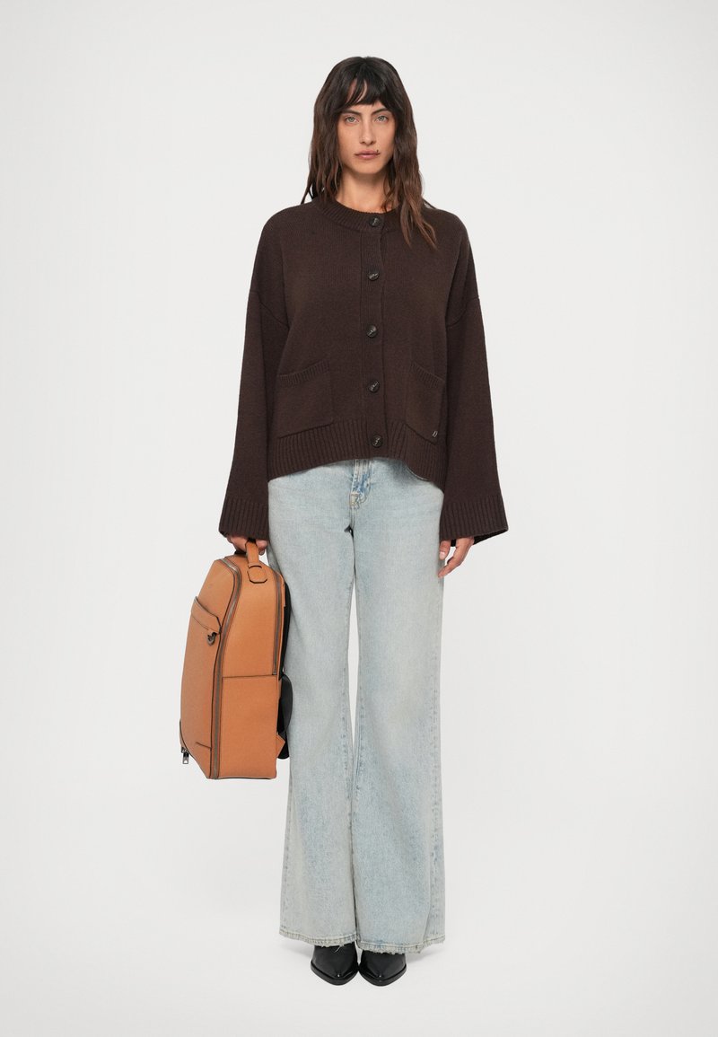 Bruine gebreide cardigan met knopen en frontzakken, gecombineerd met lichtblauwe wijde jeans en een bruine rugzak. Zwarte puntige schoenen.