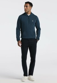 Donker teal hoodie met een kangaroo pocket, verstelbare drawstring capuchon en een klein logo op de borst. Gecombineerd met donkere broek en witte sneakers.
