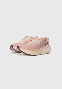 Des chaussures de sport rose clair avec un dessus texturé, des lacets crème et des accents violets sur la semelle extérieure, dotées d'un design de semelle intermédiaire rembourrée.