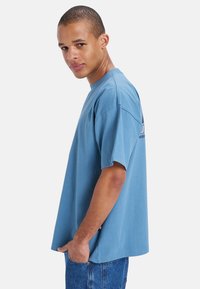 Blå bomull t-skjorte med korte ermer, rund hals, avslappet passform, og trykt logo i svart på baksiden. Brukt sammen med blå denim jeans.